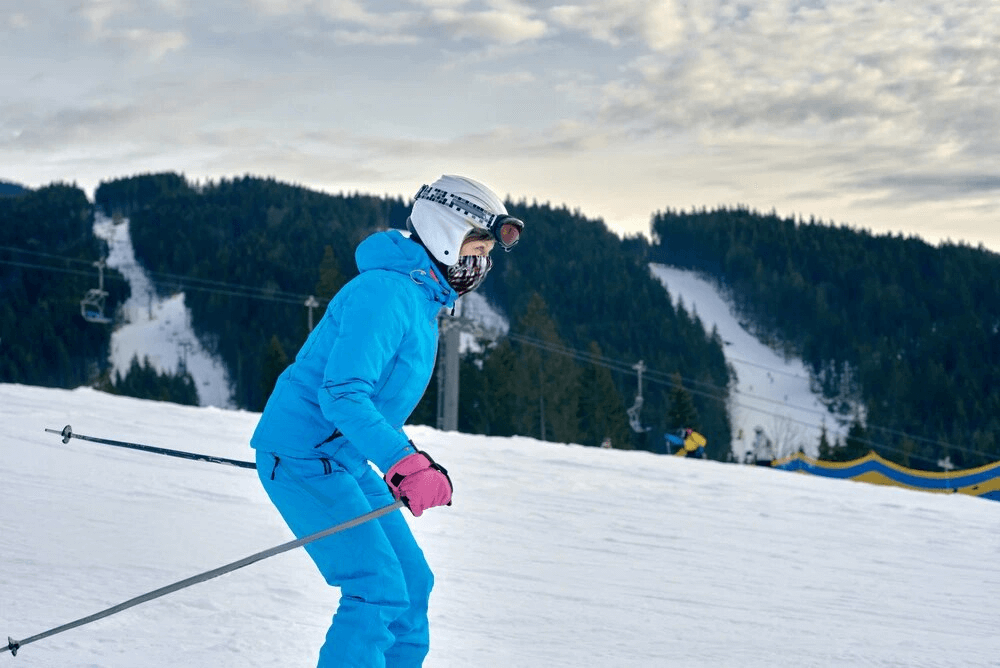 Skier in blue outfit with helmet and goggles gliding down snowy slope, scenic mountain view in the background under a cloudy sky.